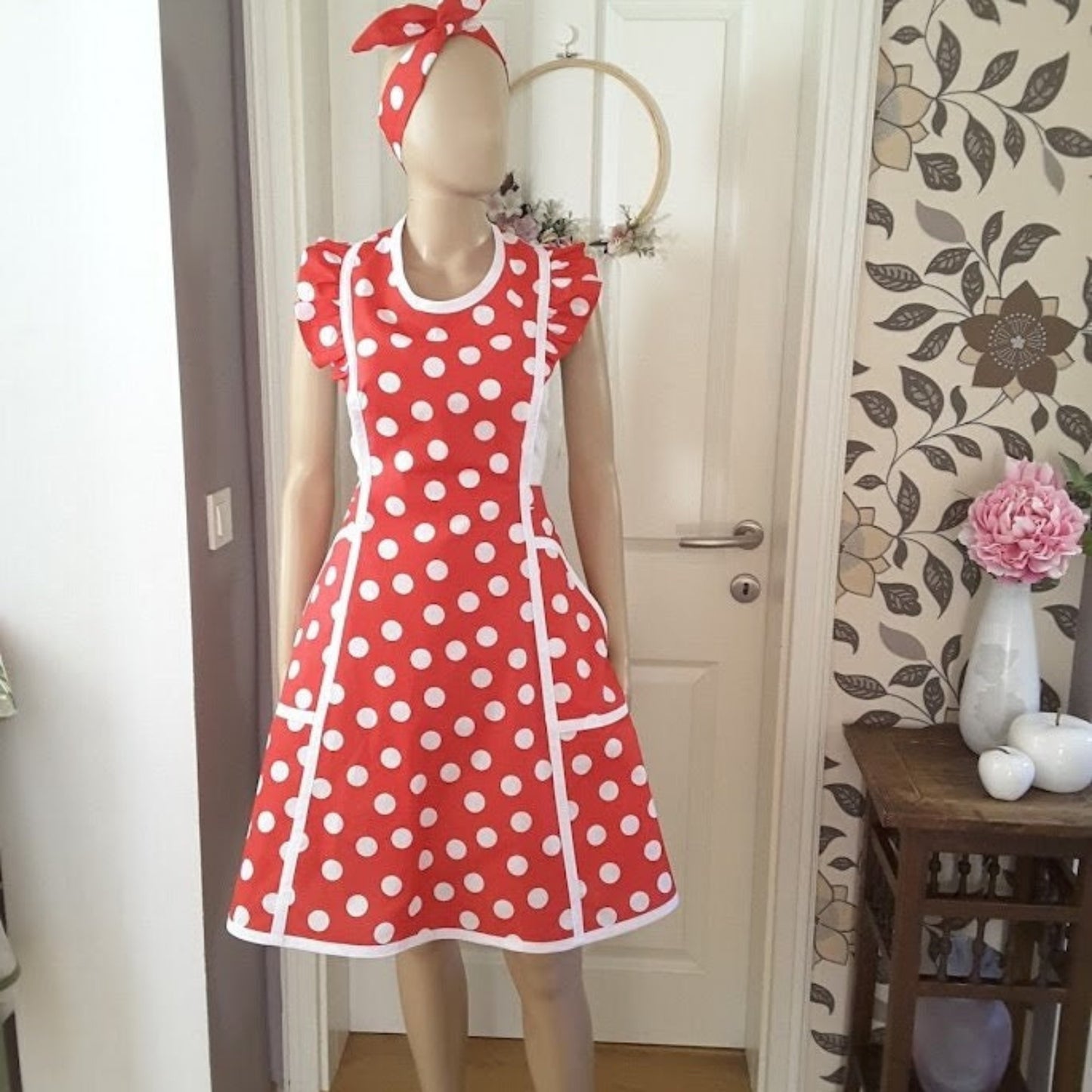 A red and white polka dot apron with white bias trim, pockets, and shoulder ruffles displayed on a mannequin. A matching headband is also visible.