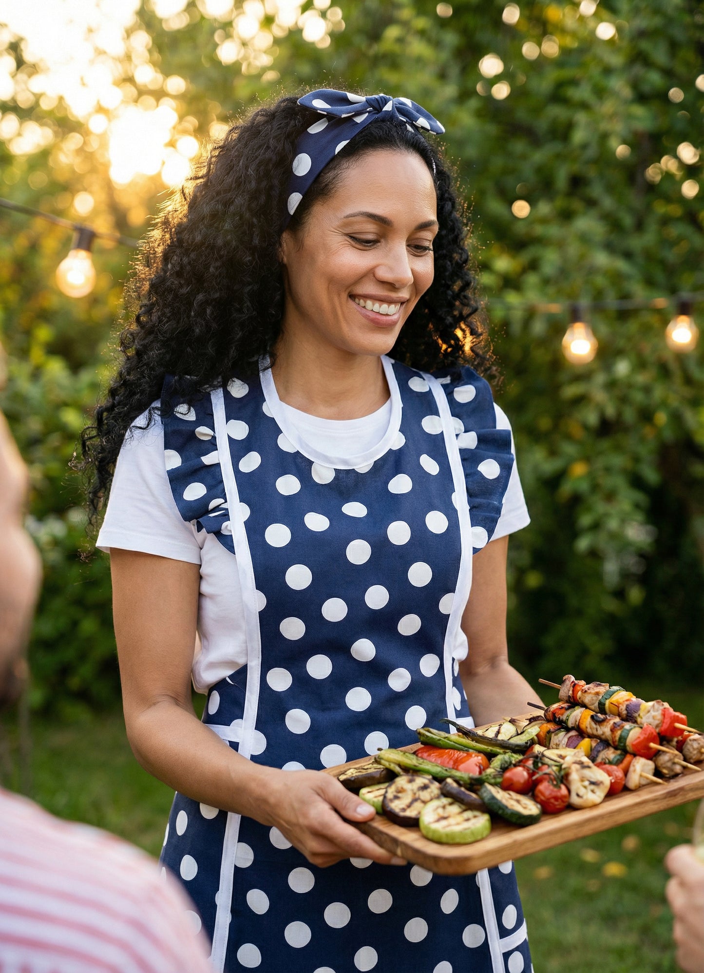 Woman in a blue polka dot retro style apron and a matching headband, holding a tray of grilled vegetables outdoors