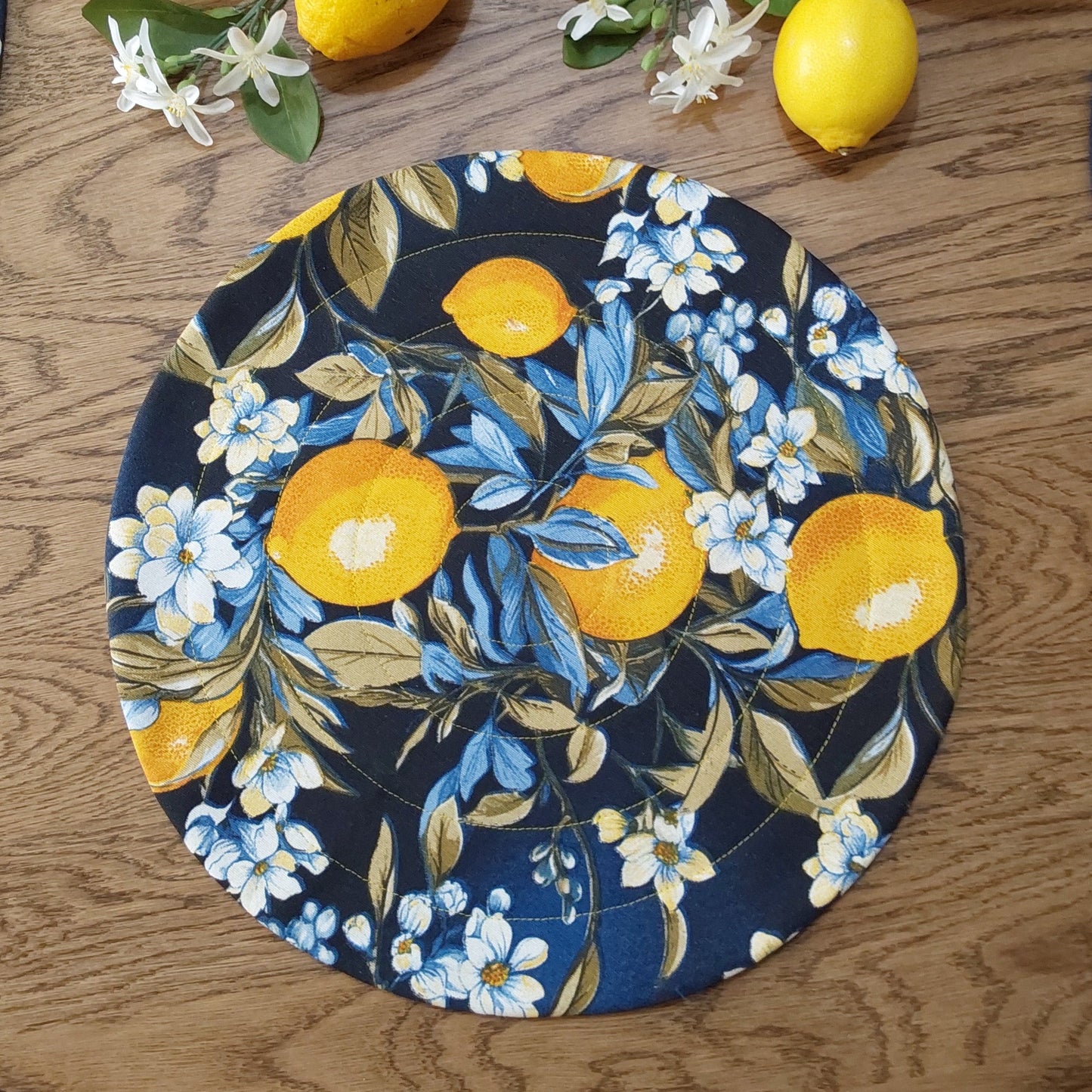 A round quilted placemat with a dark blue background featuring a bold lemon print surrounded by floral designs.