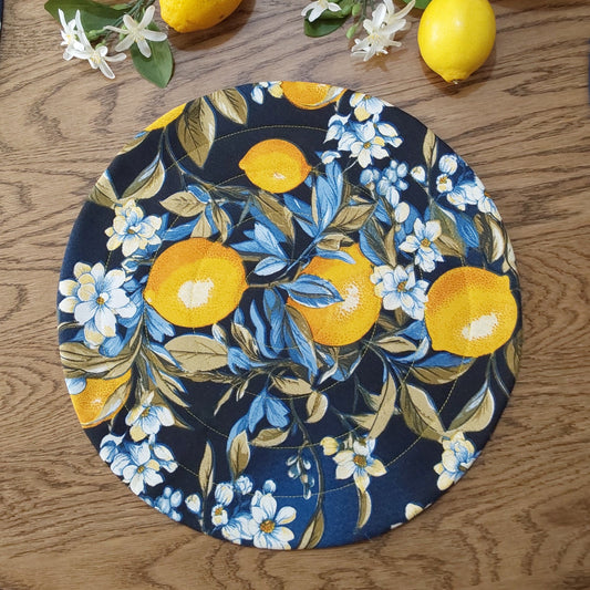 A round quilted placemat with a dark blue background featuring a bold lemon print surrounded by floral designs.