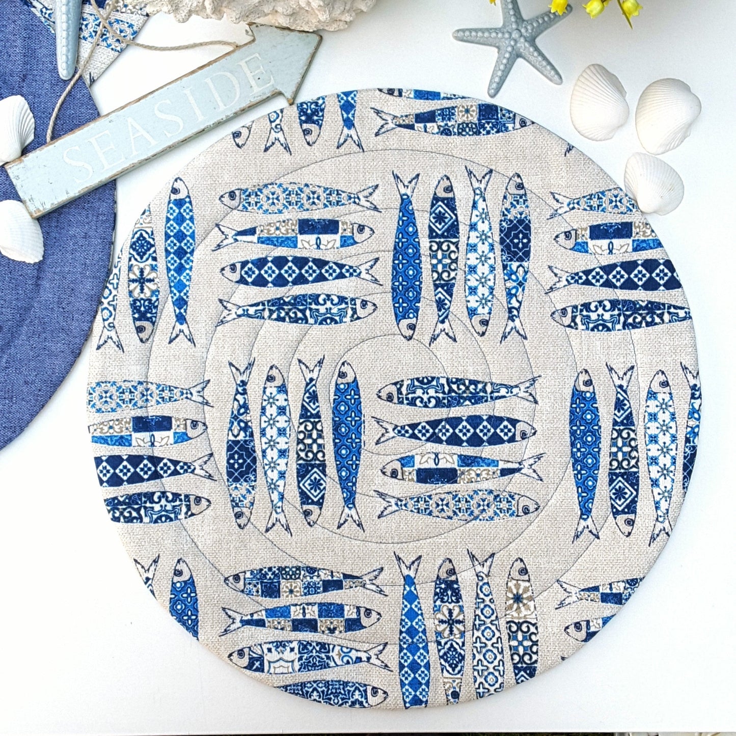 A round quilted placemat with a beige background and a blue sardines fish print