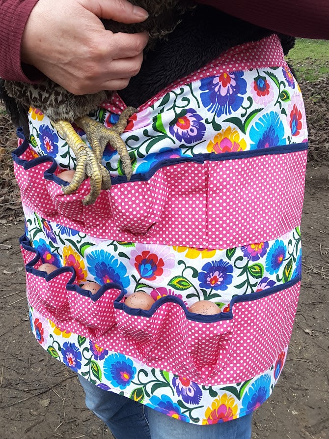 A person wearing a pink floral and polka-dot apron with multiple pockets, holding a chicken.